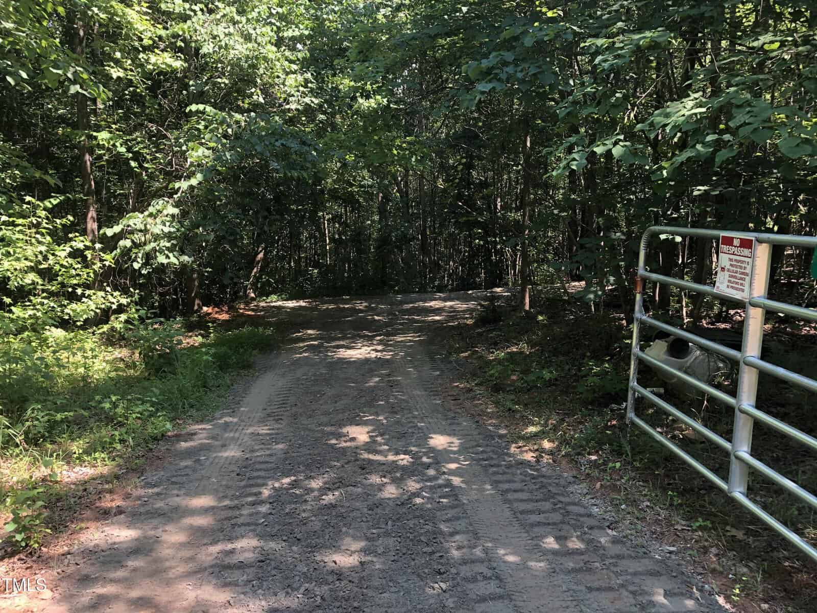 A dirt road surrounded by dense green trees curves ahead, with a metal gate and warning sign on the right side. Sunlight filters through the foliage, creating dappled shadows on the ground.