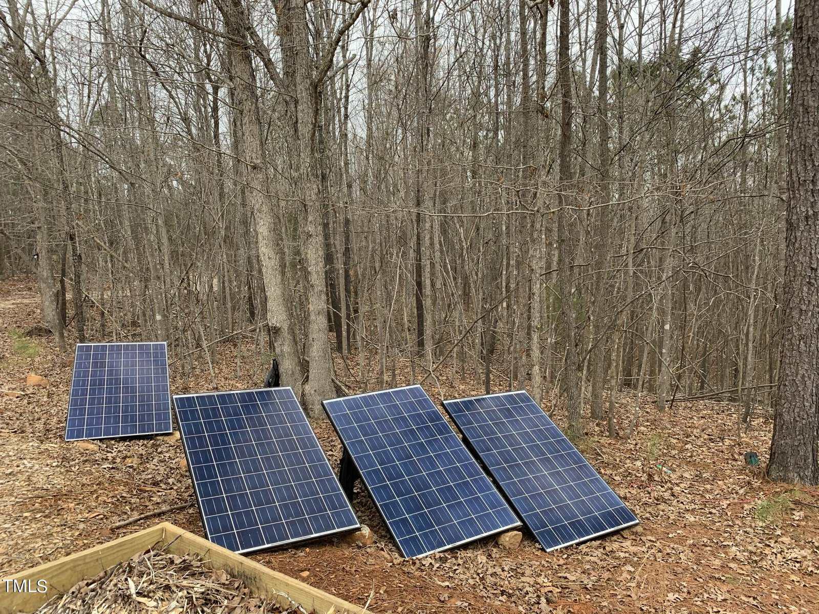 Four solar panels are set up on the ground in a wooded area with leafless trees, surrounded by fallen leaves. The sky is overcast, and there is a wooden structure partially visible in the foreground.
