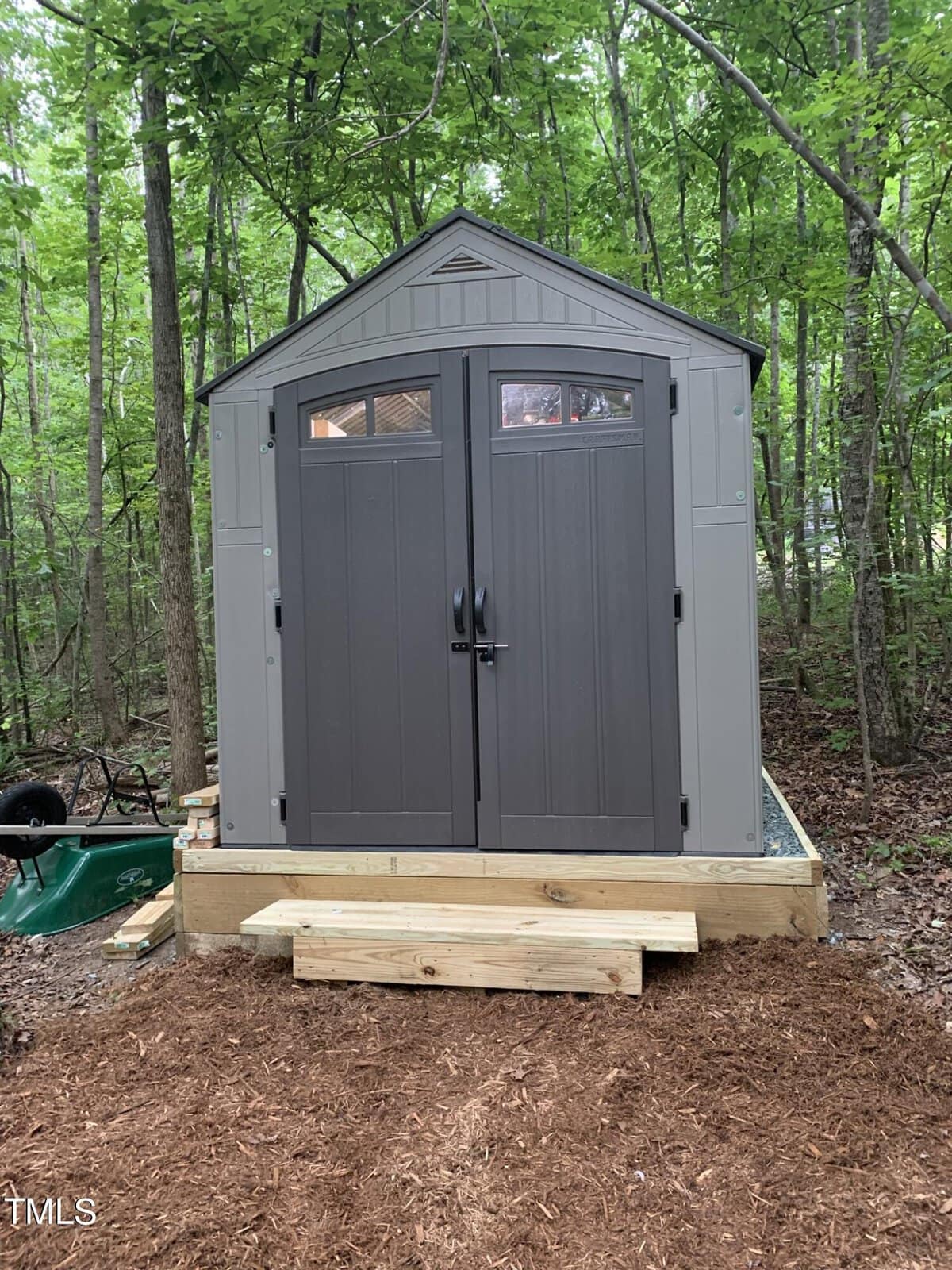 A gray plastic storage shed with double doors and small windows stands on a raised wooden platform in a wooded area, surrounded by trees and mulch. A green wheelbarrow sits to the left.