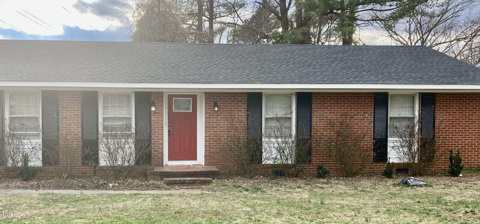 A single-story brick house with a red door and a shingled roof, located at 224 White Oak Drive, Henderson NC, is fronted by a lawn with scattered grass and bare shrubs