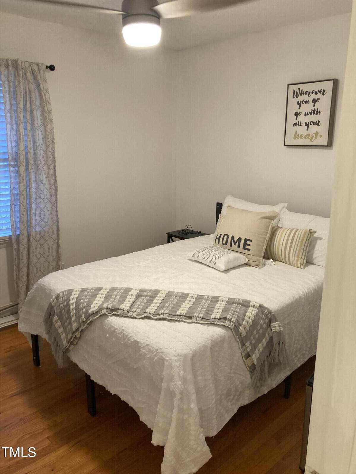 A neatly arranged bedroom with a made bed, white bedding, a decorative throw, and a pillow with the word "home" on it. on the wall, there's a framed quote, and there is a side table with a lamp next to the bed. the room is well-lit and has a simple, clean decor.