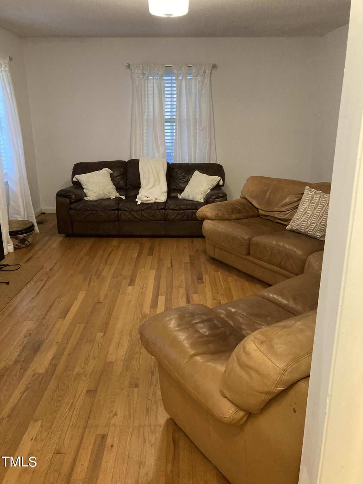 A living room featuring a brown leather sofa with white cushions, a matching armchair, and hardwood flooring. light streams in through the window with sheer curtains.