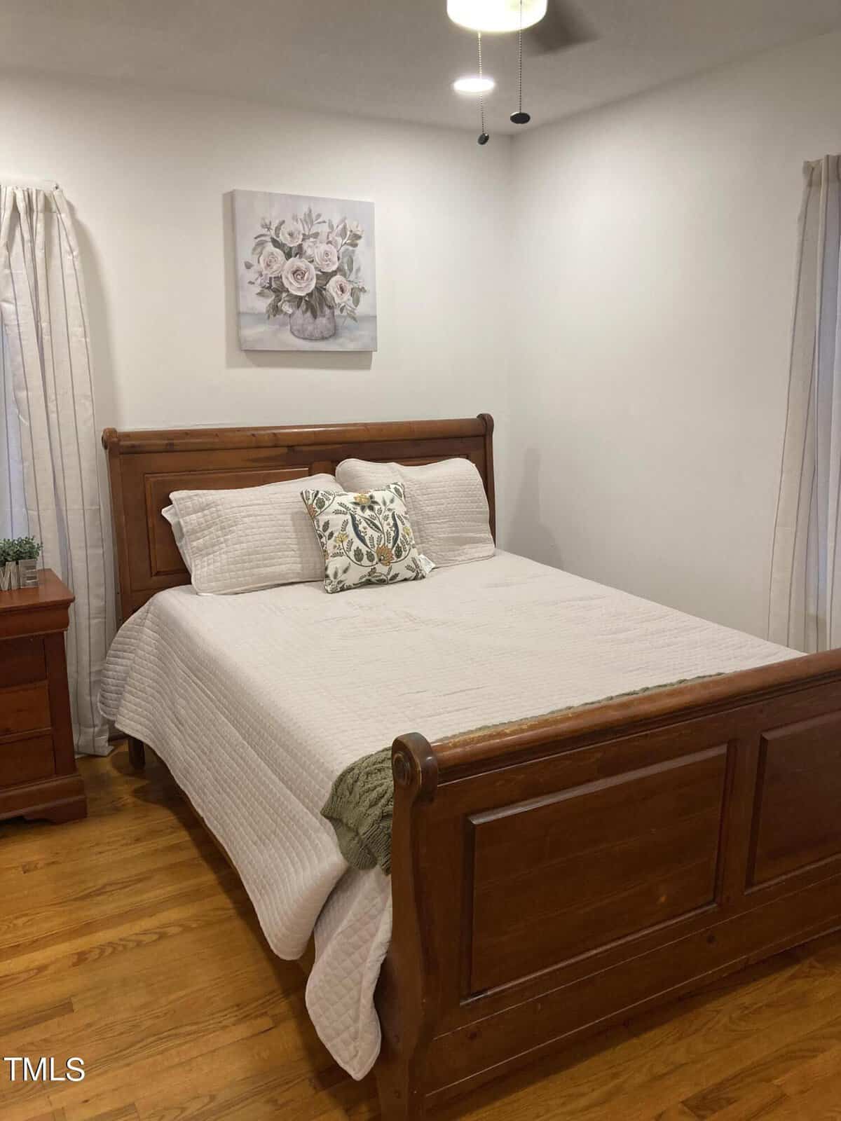 A neatly arranged bedroom with a wooden queen-size bed, white bedding, and a floral accent cushion, flanked by a matching wooden nightstand, with a decorative plant and a wall-mounted artwork above the headboard.