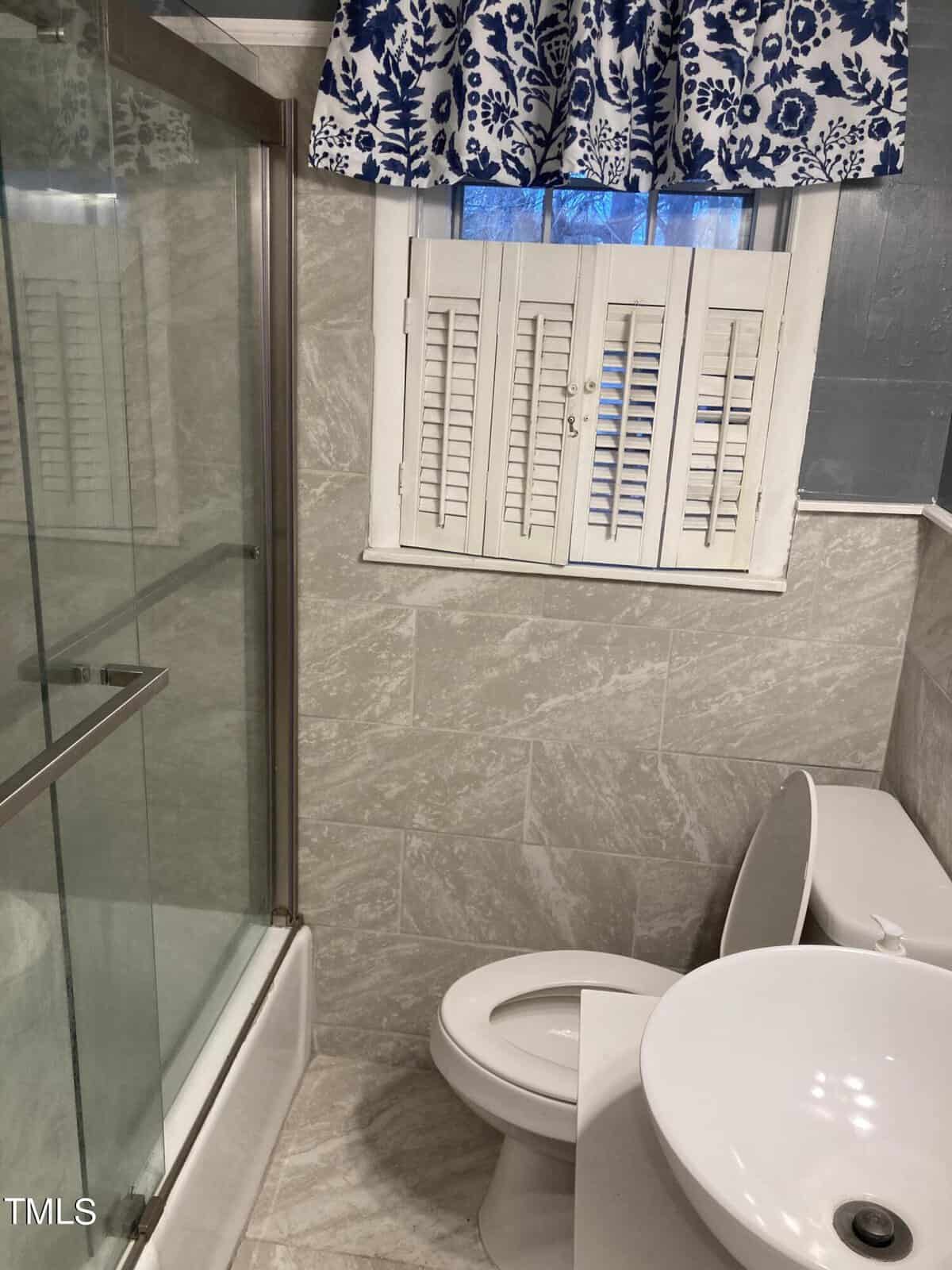A bathroom with a glass shower enclosure to the left, a toilet in the center, and a window with closed white shutters and a blue-patterned valance above.