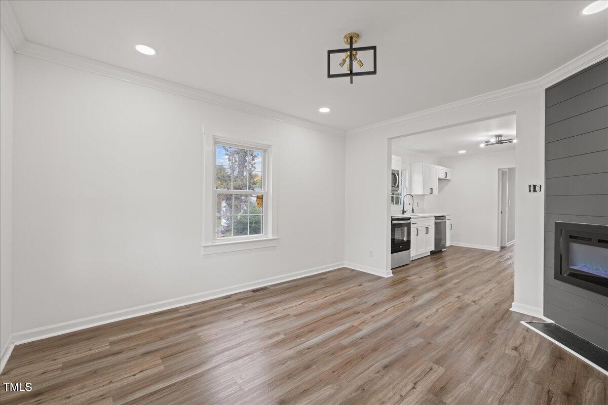 An empty room with light hardwood flooring, white walls, and a fireplace. a peek into an adjoining kitchen reveals white cabinets and black countertops. two windows provide natural light.