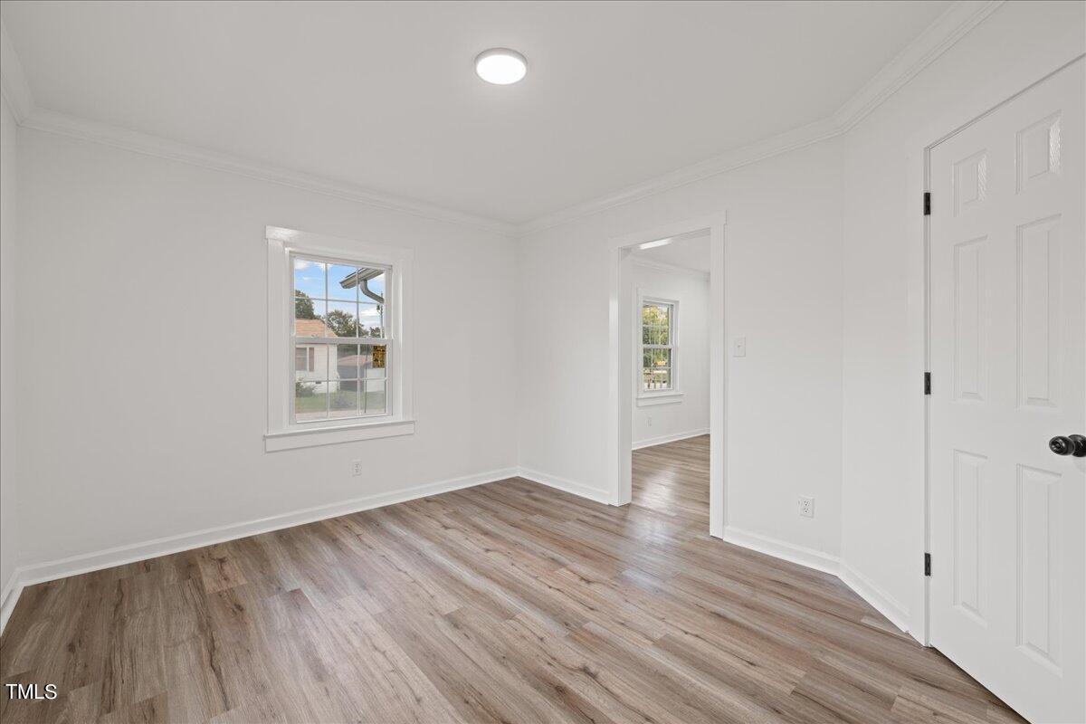 An empty room with white walls and light brown wooden flooring, featuring a single window and an open door. the room is brightly lit with a central ceiling light.