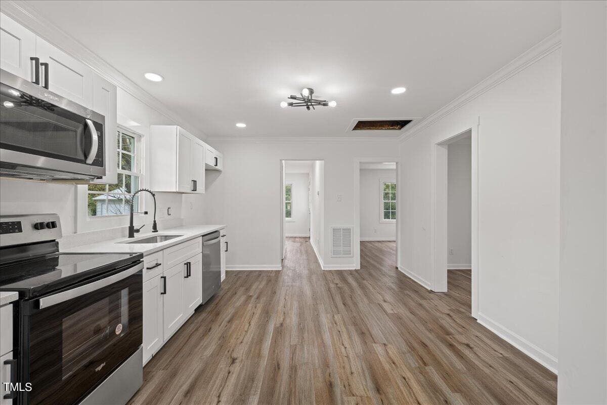 A modern kitchen with white cabinetry, stainless steel appliances, and a dark countertop. the room features light wooden flooring, multiple doorways, and a centrally located ceiling light.