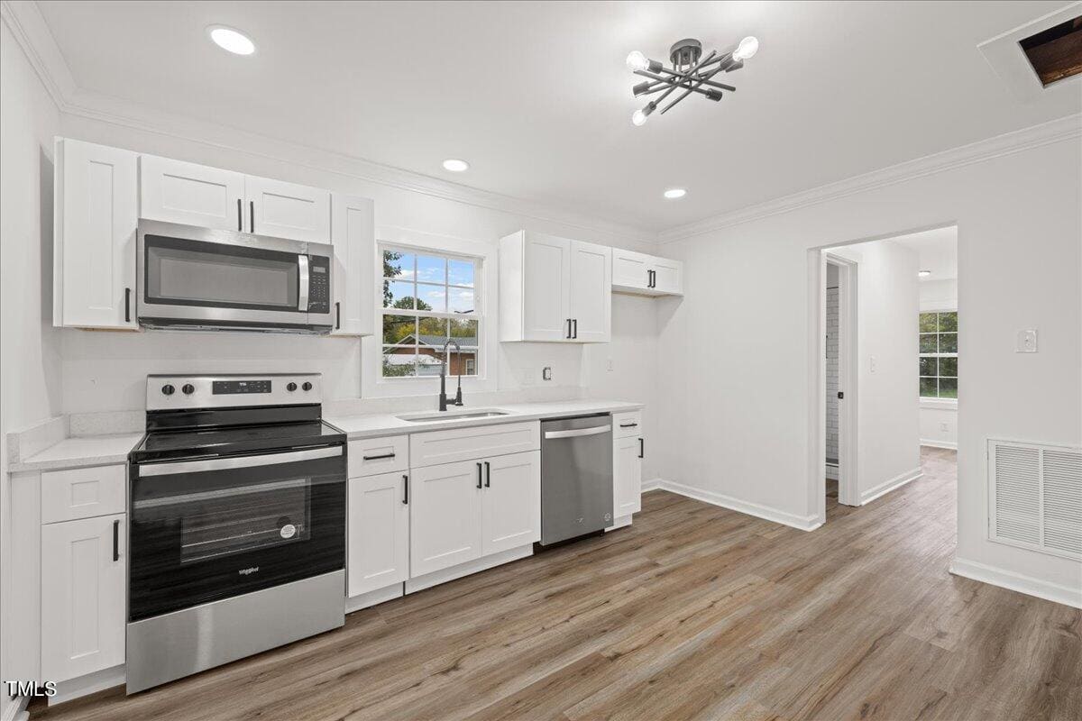A modern kitchen with white cabinets, stainless steel appliances, and wooden flooring. the room is well-lit with recessed lighting and a ceiling fan, featuring a view of greenery through the window.
