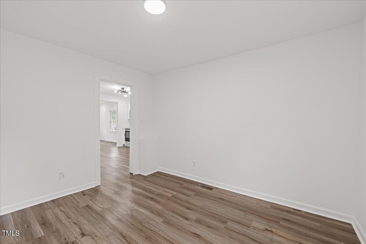 An empty room with white walls and wooden flooring, featuring a single ceiling light and an open doorway leading to another room.