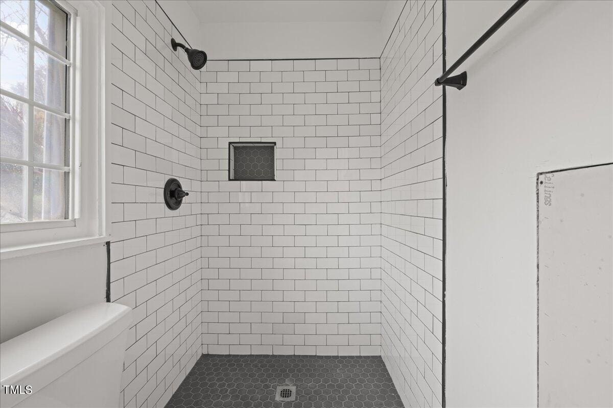 Modern bathroom with white subway tiles on walls, a corner shower with black fixtures, and gray hexagonal floor tiles. there's a small window, and a partial view of a white bathtub.