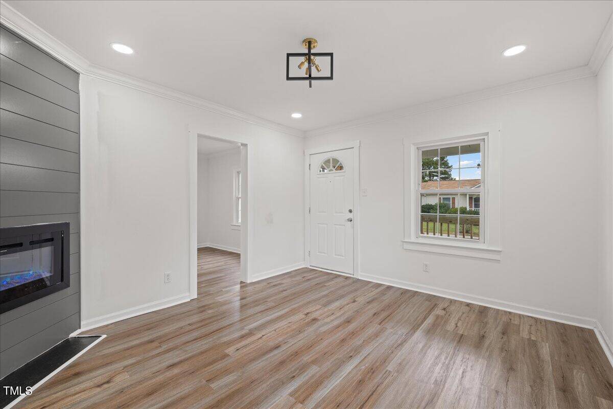 An empty room featuring gray hardwood floors, a dark gray accent wall with an electric fireplace, white walls, and a white front door with a semi-circular window. there is a modern black and gold light fixture hanging from the ceiling.
