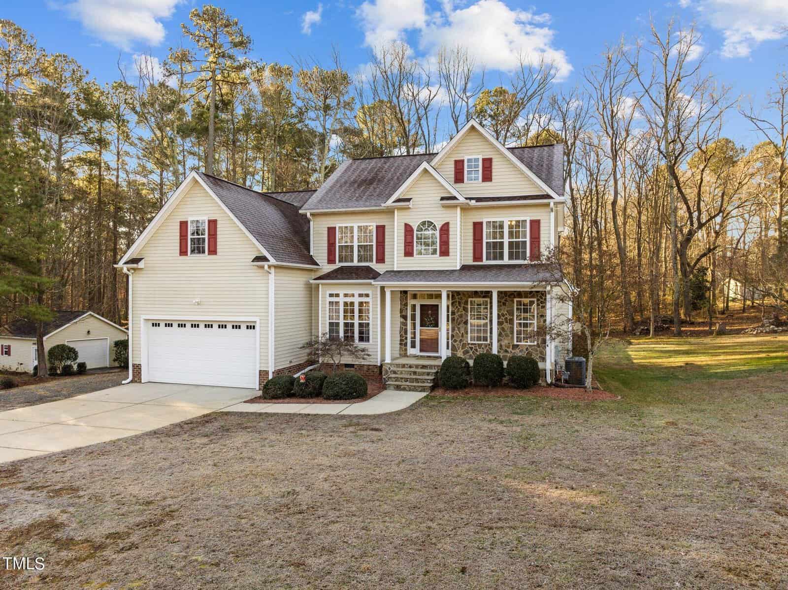 A two-story suburban house with a beige exterior and red shutters, surrounded by a grassy yard with trees. The house has a double garage and a small front porch. The sky is blue with a few clouds.