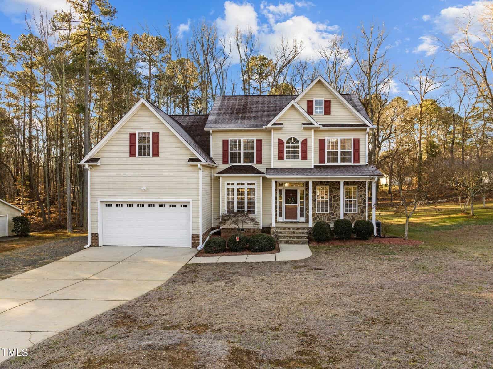 Nestled in Wake Forest on 3696 Morgan Circle, this charming two-story house boasts beige siding and brick accents, with red shutters enhancing its charm. A two-car garage and wide driveway complete the picturesque scene against a backdrop of trees under a clear blue sky.