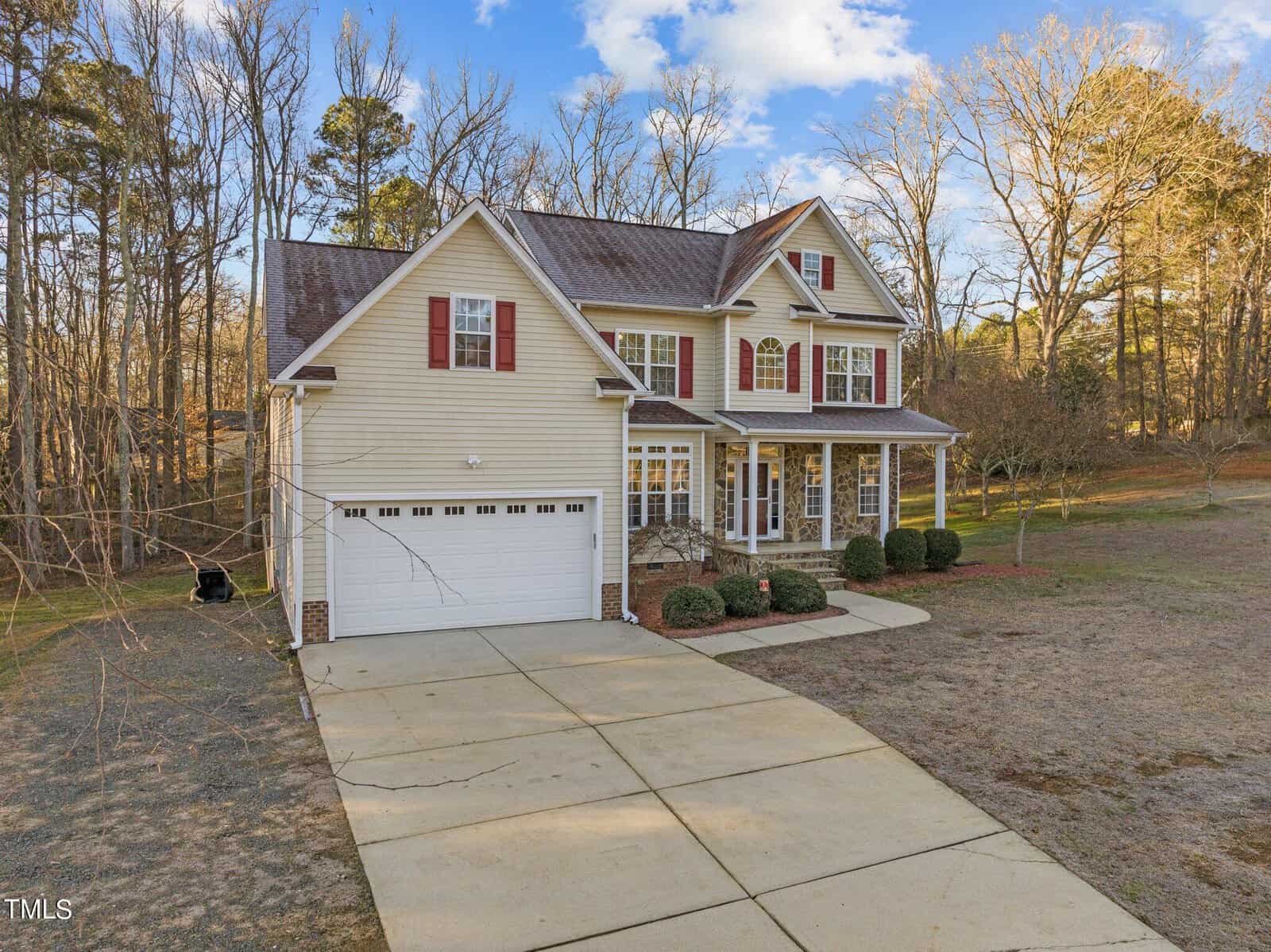 A two-story, beige house with a double garage, red shutters, and a front porch with columns. It is set in a wooded area with bare trees and a large concrete driveway, under a partly cloudy sky.