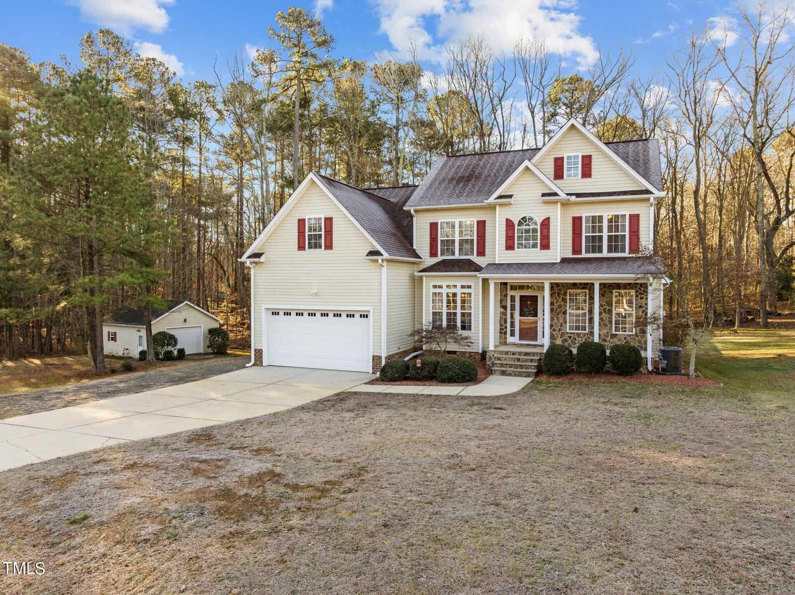 A two-story house with beige siding and red shutters, featuring a two-car garage and a small porch. The front yard is mostly bare, and the property is surrounded by tall trees under a blue sky with scattered clouds.