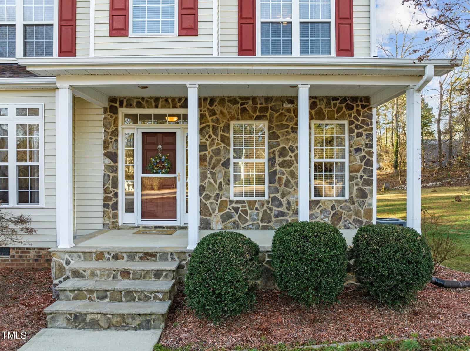 The image shows the front entrance of a two-story house with stone and beige siding, featuring a small porch with white pillars, a wreath on the door, and manicured bushes lining the walkway.