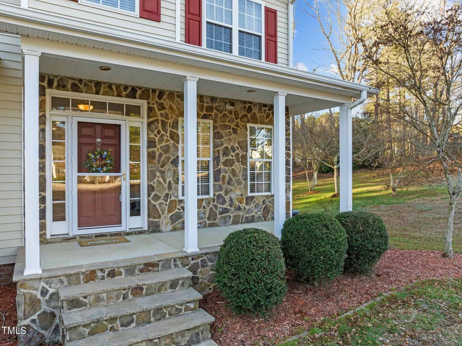 Front view of a house with a stone facade, a red door decorated with a wreath, white columns, and a covered entryway. The yard is landscaped with shrubs and mulch, and trees are visible in the background.