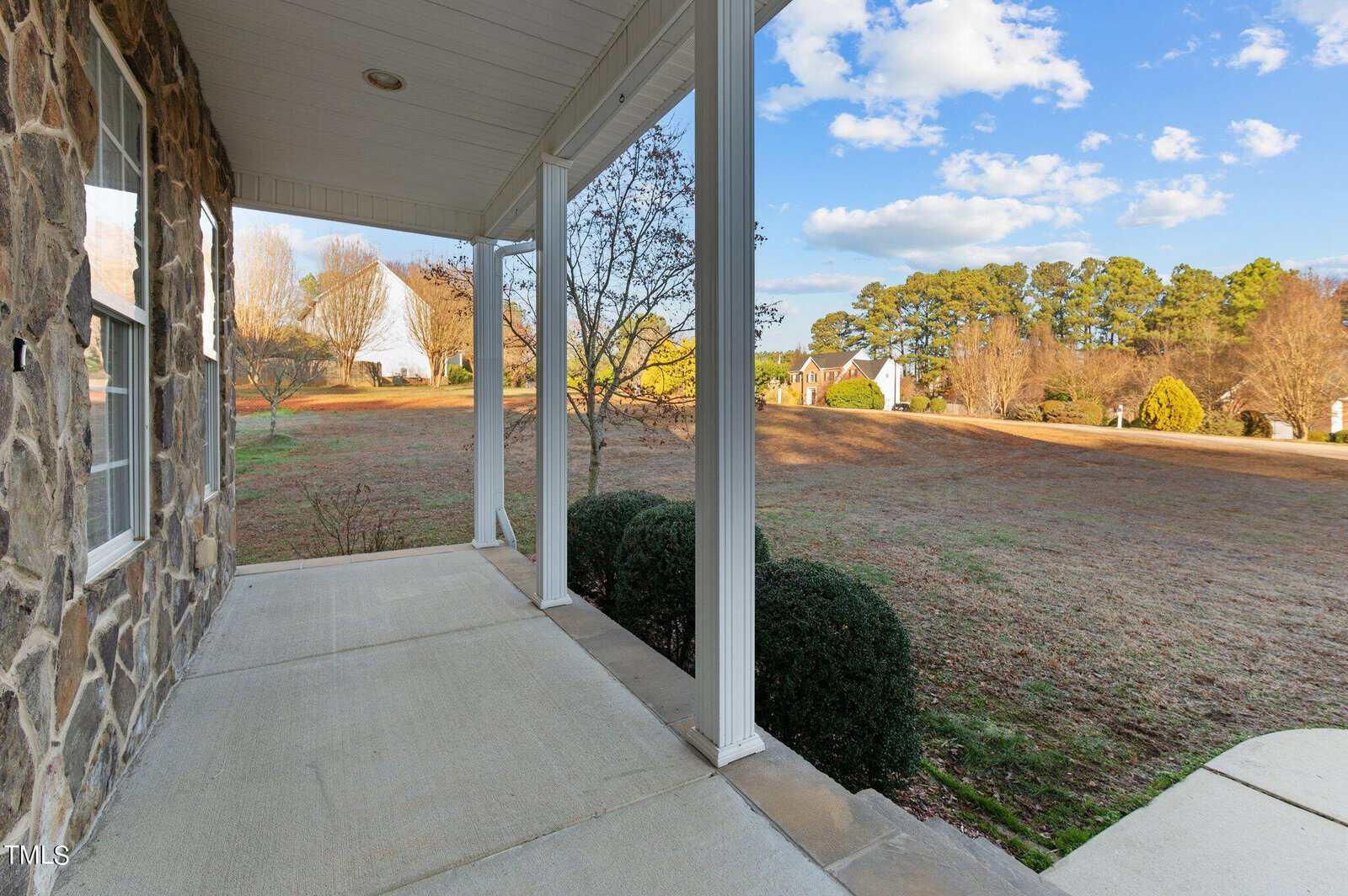 Stone porch with a view of a spacious grassy yard dotted with trees and houses in the distance. A clear blue sky with scattered clouds adds to the peaceful suburban scene.