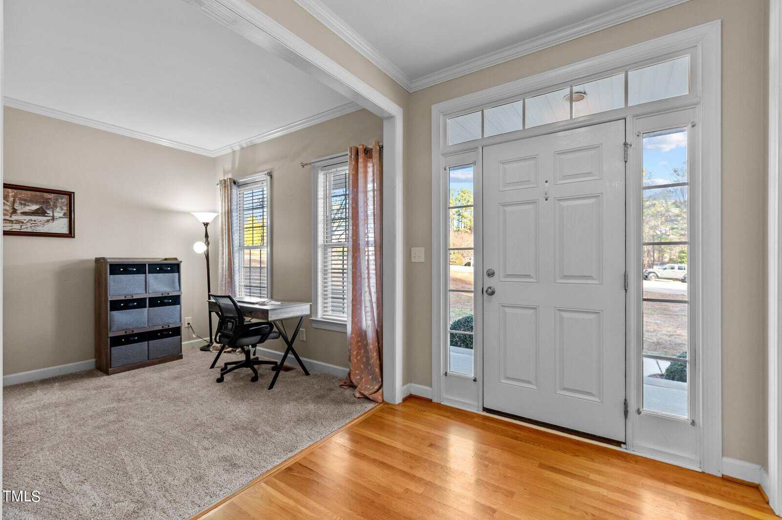 A bright room with hardwood floors and a carpeted area features a desk and chair by two windows with curtains. There's a white front door with glass panels on the right. A wood chest with drawers is on the left, and a framed picture hangs on the wall.