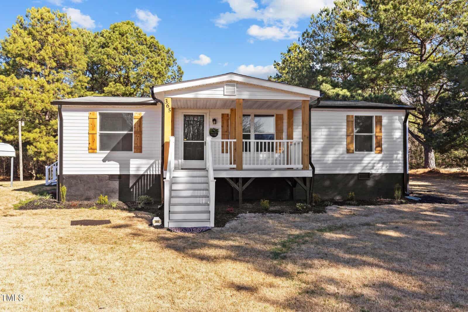 A single-story manufactured home with a white exterior and gray trim, featuring a small front porch with steps leading up to the entrance, surrounded by a natural landscape with pine trees.