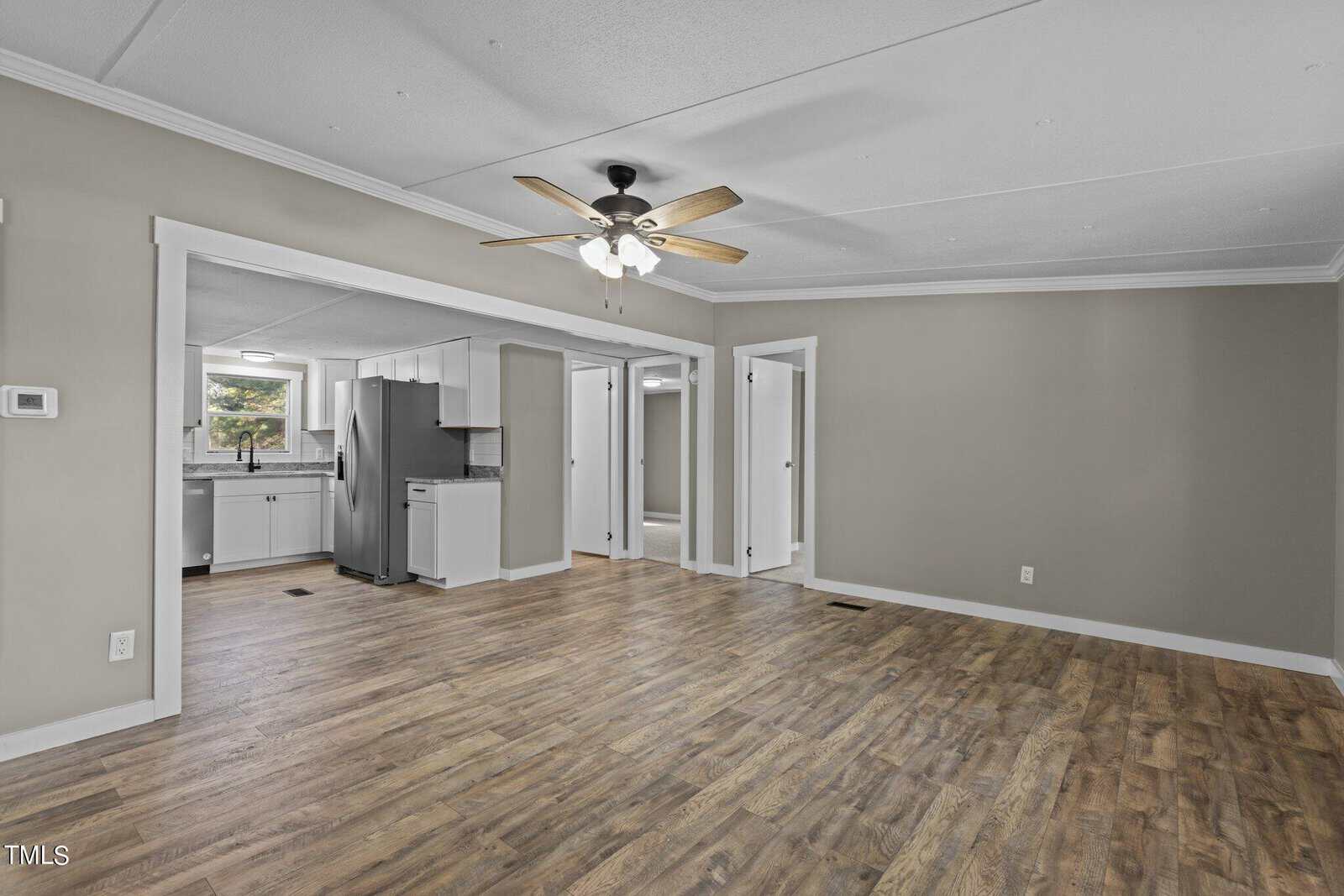 An empty room with wooden flooring, featuring a ceiling fan, leading to an adjacent kitchen area with modern appliances, and multiple doors along the wall.