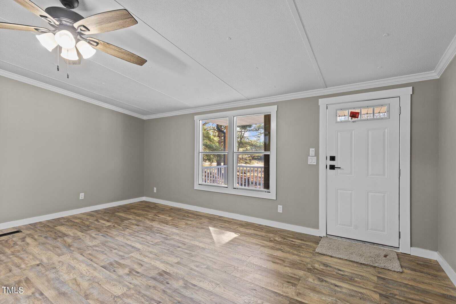An empty room featuring light gray walls, wood laminate flooring, a ceiling fan, and a sliding window with a view of trees. a white door with a deadbolt is on the right and there is a beige doormat in front of it.