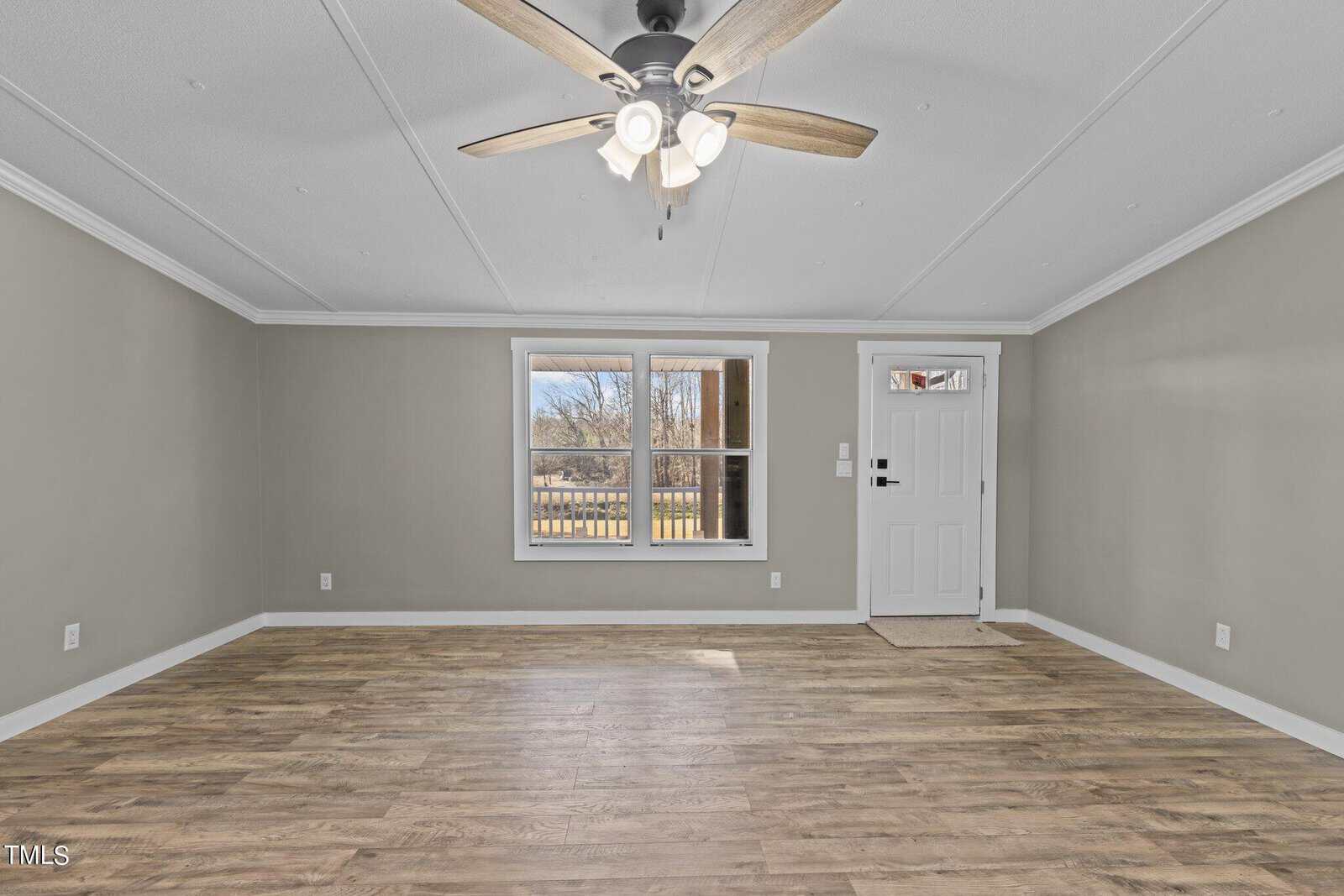 An empty room with wooden flooring, white walls, a ceiling fan, and a window showing an outside view. a white door is visible on the right.