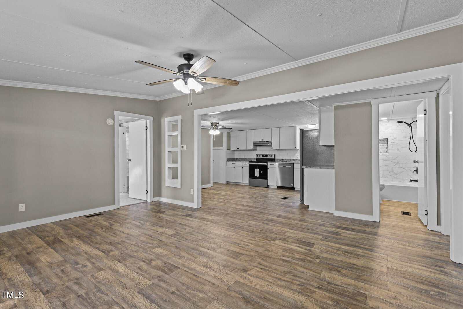 An empty, modern room featuring wood laminate flooring, a ceiling fan, and an open-plan layout leading into a kitchen with stainless steel appliances and white cabinetry.
