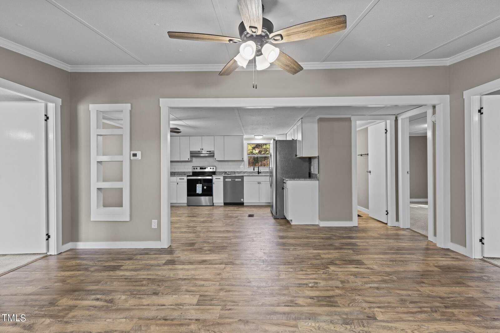 An open-concept interior space featuring a combined living and dining area with hardwood flooring, a ceiling fan above, white walls, and an adjacent kitchen with modern appliances. the layout includes multiple entryways and built-in shelving or cabinetry on the left.