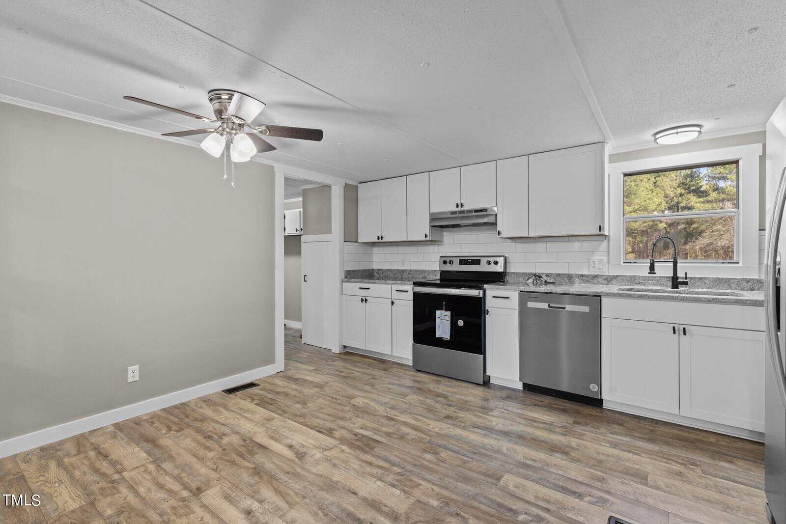 A modern kitchen with white cabinetry, stainless steel appliances, and light wood flooring, adjacent to a living area with a ceiling fan.