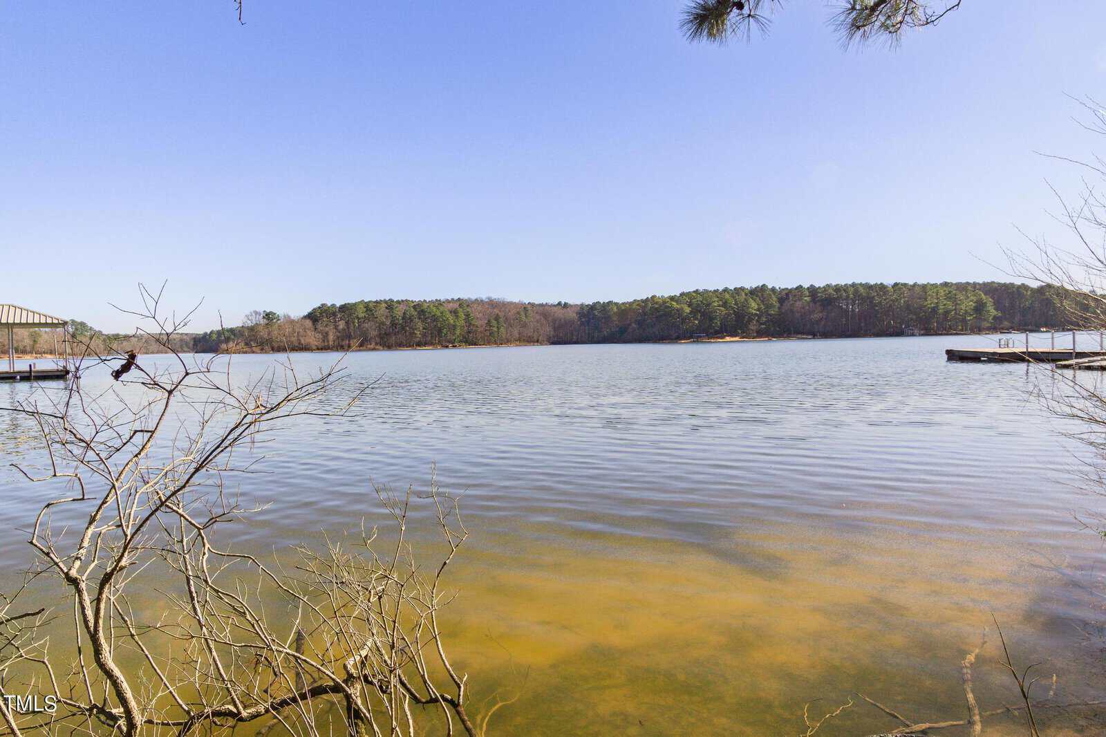 A tranquil lake scene with clear blue skies and a distant tree-lined shore. The water is calm, and leafless branches extend from the bottom left. Two docks are visible on either side of the image.