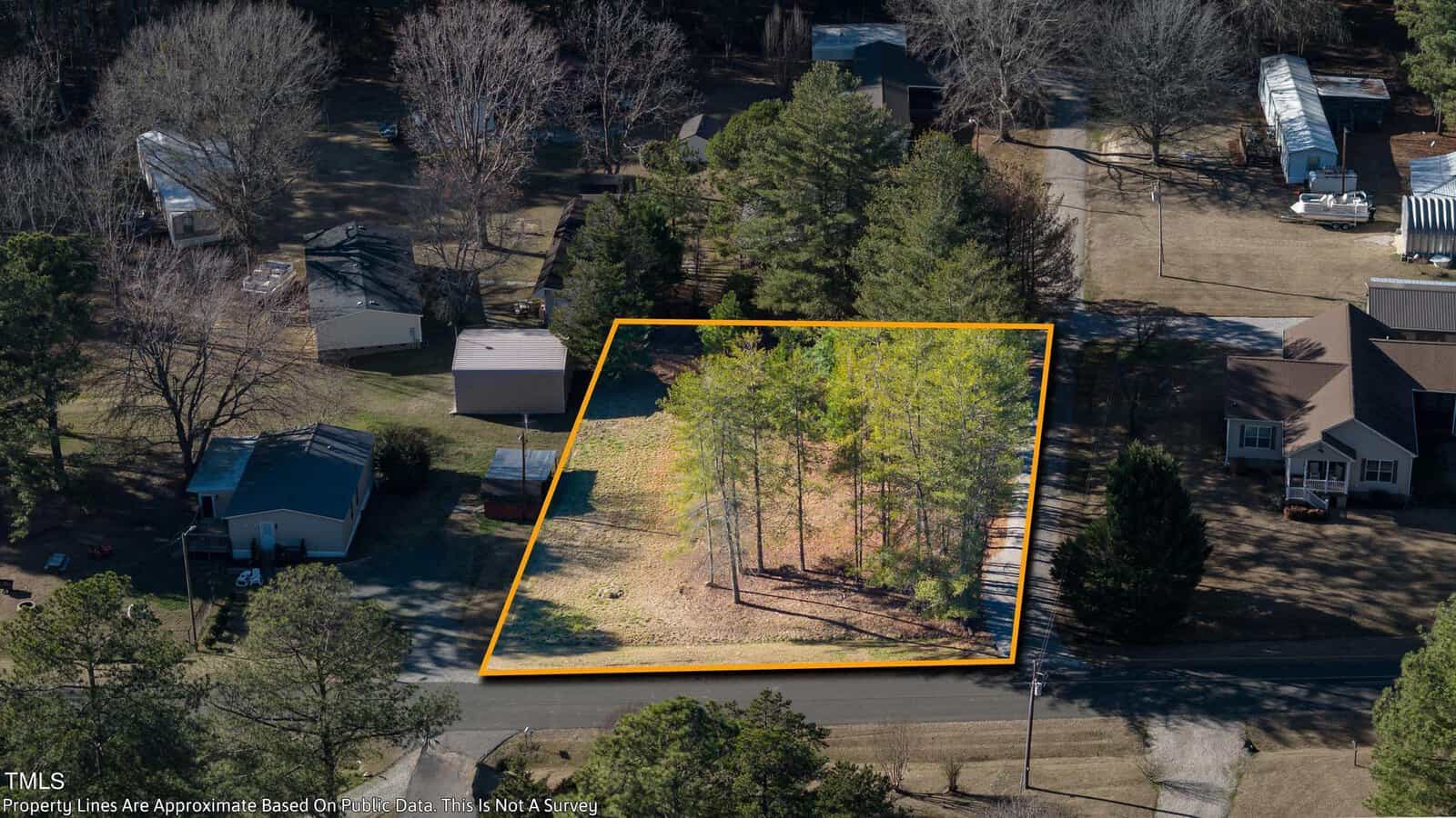 Aerial view of a wooded lot outlined in yellow, featuring a few trees and surrounded by residential houses. The sun casts shadows from the trees onto the lawn, and a street borders the property in the foreground.