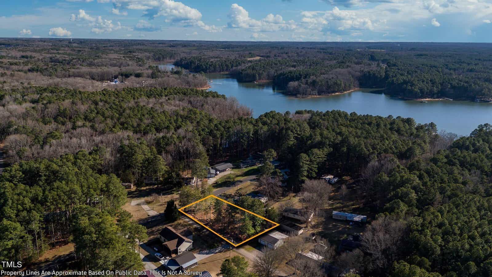 Aerial view of a forested area near a large lake. A small parcel of land is outlined in yellow, surrounded by trees and a few buildings. The sky is partly cloudy, and the landscape features dense woodland and water.