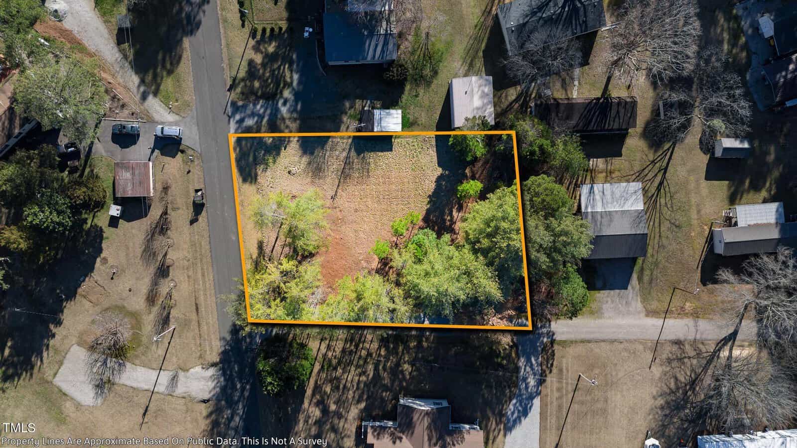 Aerial view of a rectangular property outlined in yellow. The area contains a few trees and a small structure. Nearby properties and surrounding streets are visible, with shadows indicating sunlight.