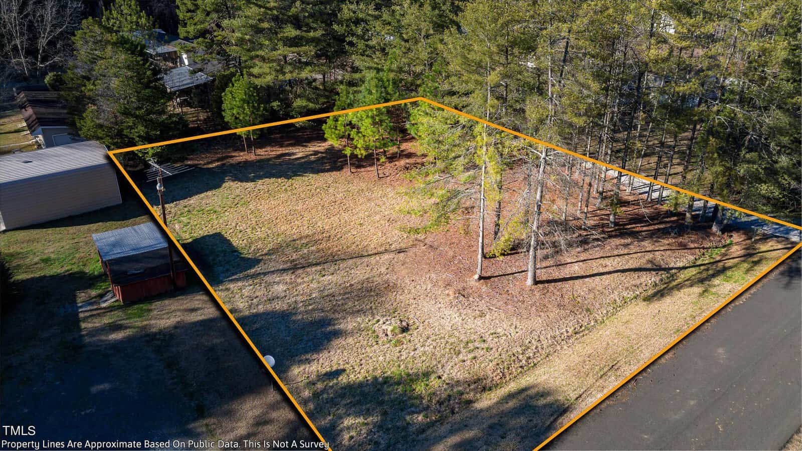 Aerial view of a rectangular plot of land bordered by orange lines. The lot has grass and a few trees, surrounded by other wooded areas and a small structure to the left. The corner of a road is visible at the bottom right.