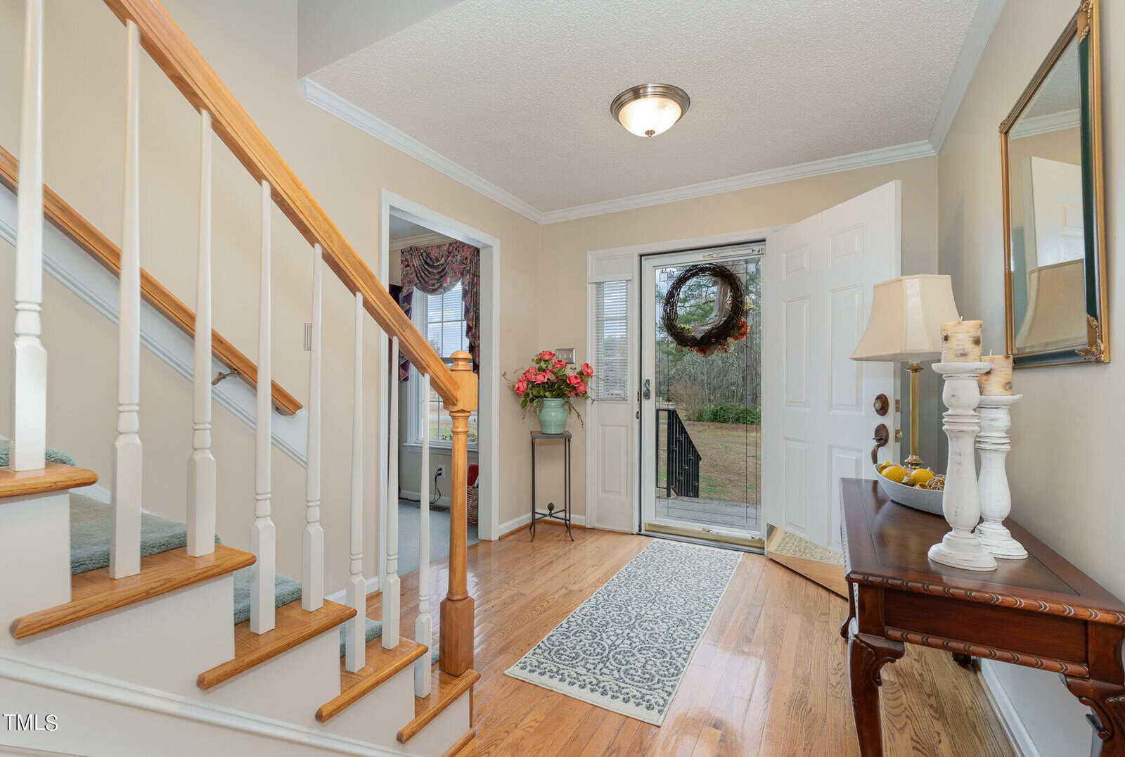 A welcoming foyer with wooden stairs, a small table holding a lamp and a bowl of lemons, and an open door adorned with a wreath. A floral arrangement stands on a pedestal. Light wooden flooring and beige walls complete the cozy look.
