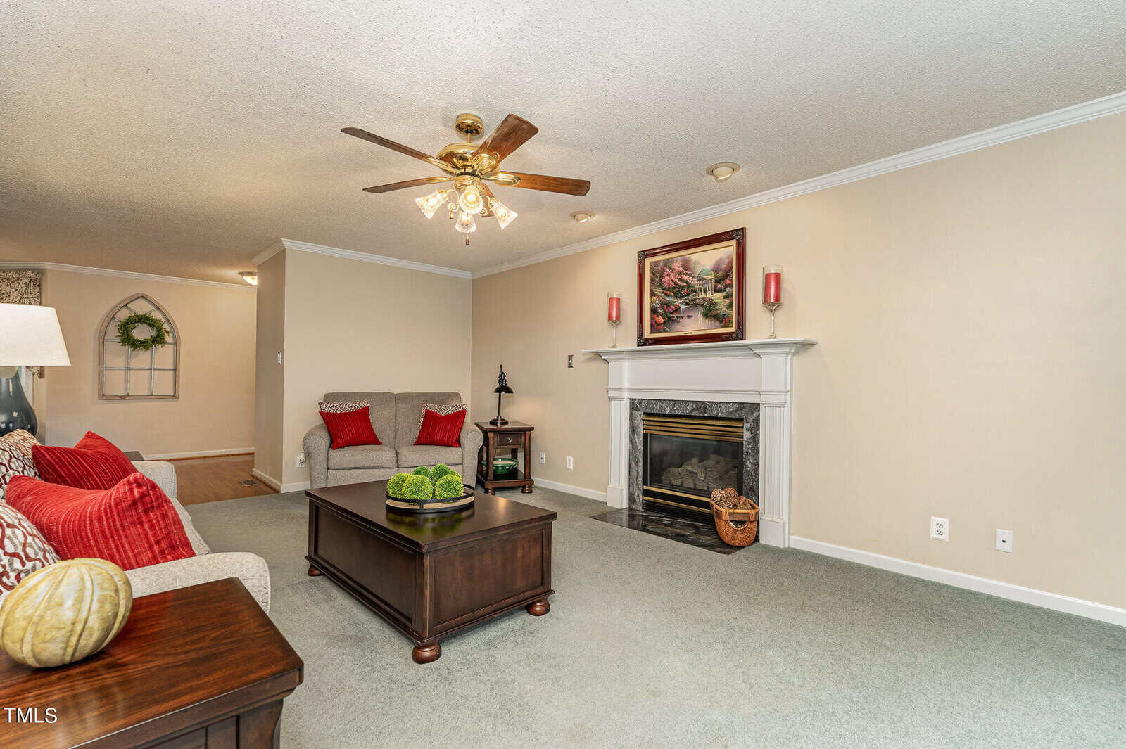A cozy living room with a ceiling fan and fireplace. The room features a gray sofa adorned with red and gray pillows, a wooden coffee table with decorative items, and a framed painting above the mantel. The carpet is a soft green color.