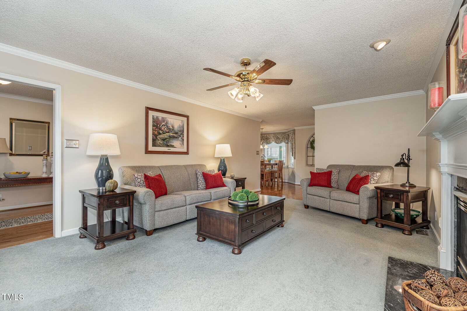 A cozy living room with two gray sofas adorned with red pillows. There's a central wooden coffee table, lamps, and a ceiling fan. A painting hangs on the wall, and a fireplace is in the corner. The room is warmly lit and inviting.