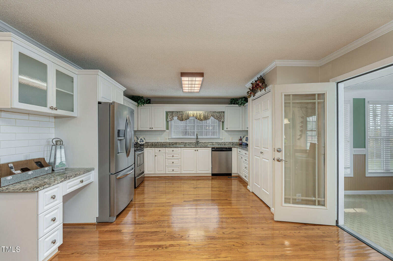 A spacious kitchen with white cabinets, granite countertops, and stainless steel appliances, including a fridge and dishwasher. There's a tile backsplash, hardwood floors, and a decorative border above the cabinets. Glass doors open to an adjacent room.