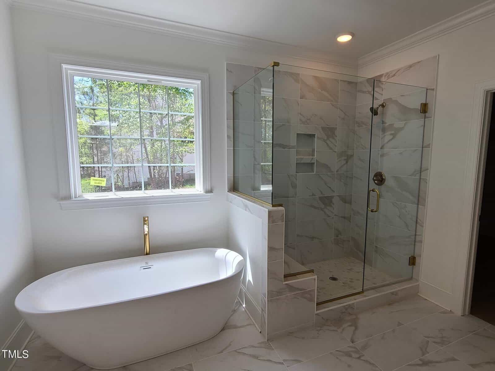 A modern bathroom with a freestanding white bathtub next to a large window. Adjacent is a glass-enclosed shower with marble tiles and gold fixtures. The room has a bright, airy feel with natural light streaming in.