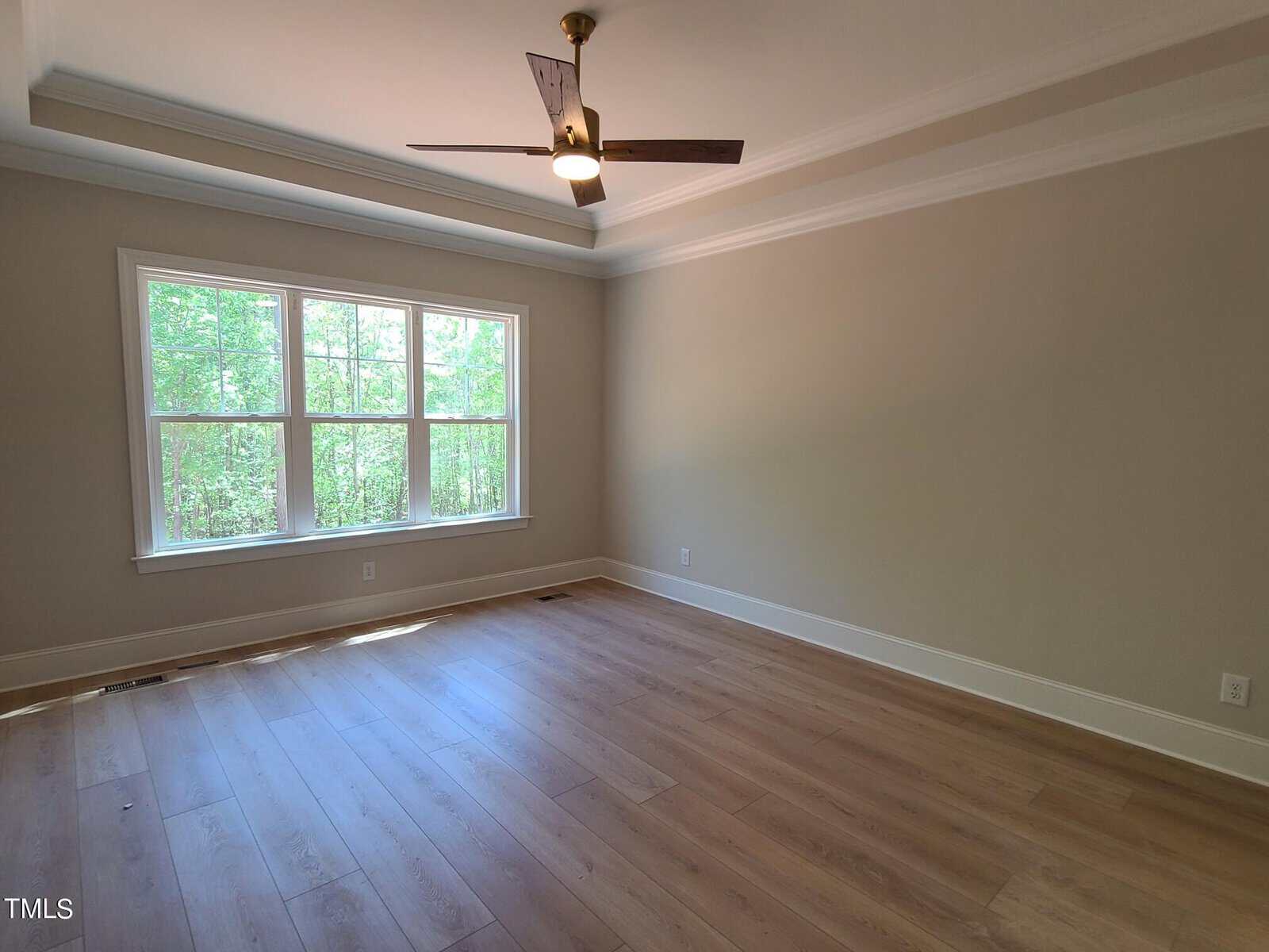 An empty room with light wood flooring and beige walls features a large window overlooking greenery. The ceiling has a fan with a light fixture. Natural light fills the space, enhancing the room's airy feel.