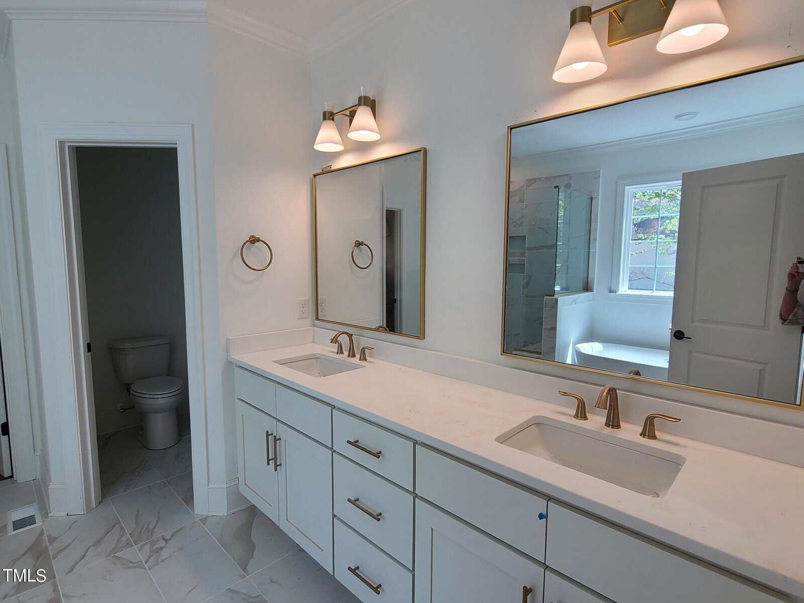 Modern bathroom with double sinks, white cabinetry, and gold fixtures. Large mirrors and twin light fixtures above each sink. A separate toilet area is visible to the left, and a window next to a bathtub is in the background.