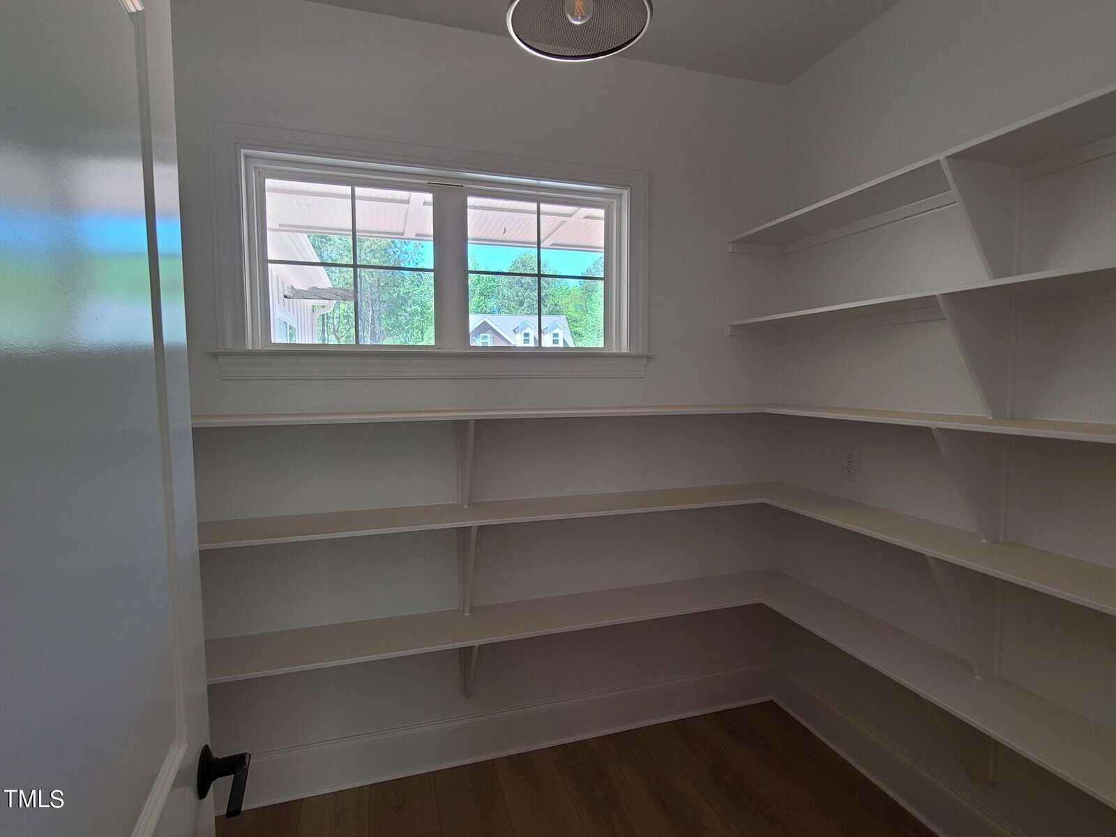 A small, empty pantry with white walls and corner shelving. There is a window with a view of the outside, bringing in natural light. The floor is wooden, and a pendant light hangs from the ceiling.