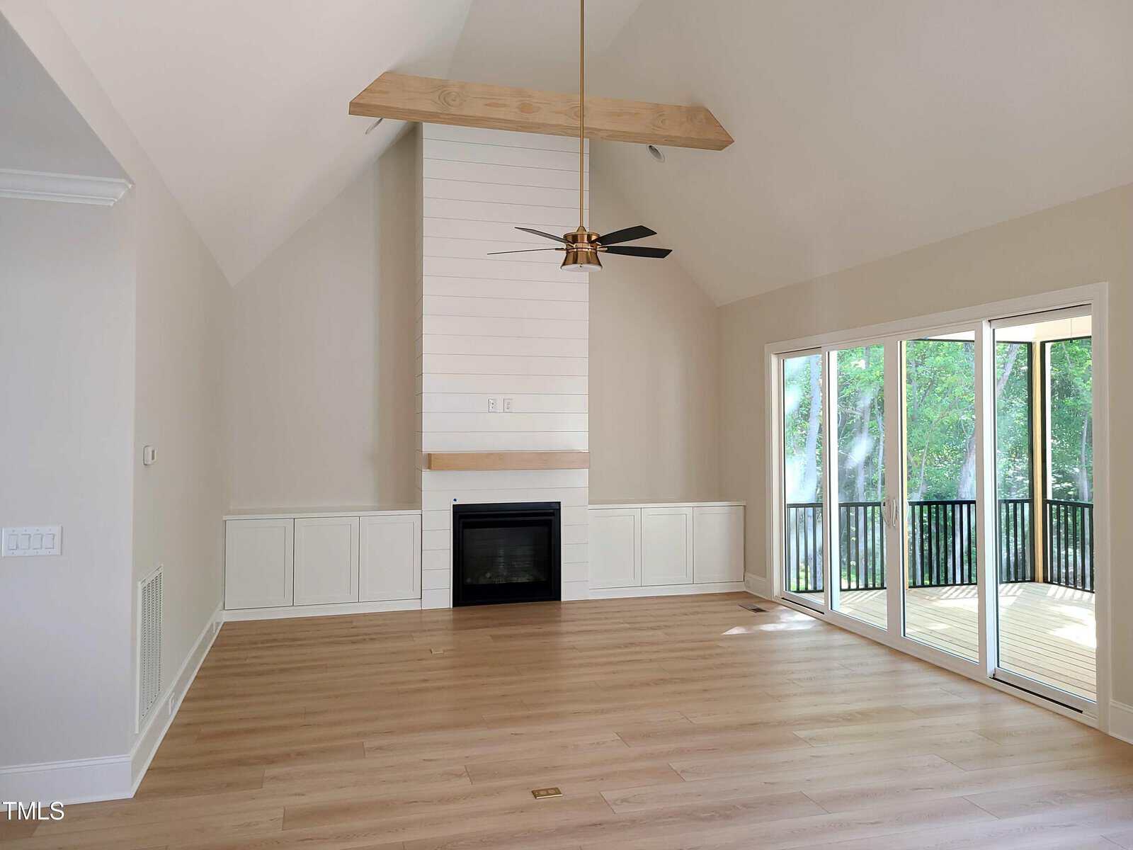 A modern, empty living room with light wood flooring, a high vaulted ceiling, and a ceiling fan with wooden beams. The room features a built-in fireplace with white shiplap paneling and large sliding glass doors opening to a deck with greenery outside.