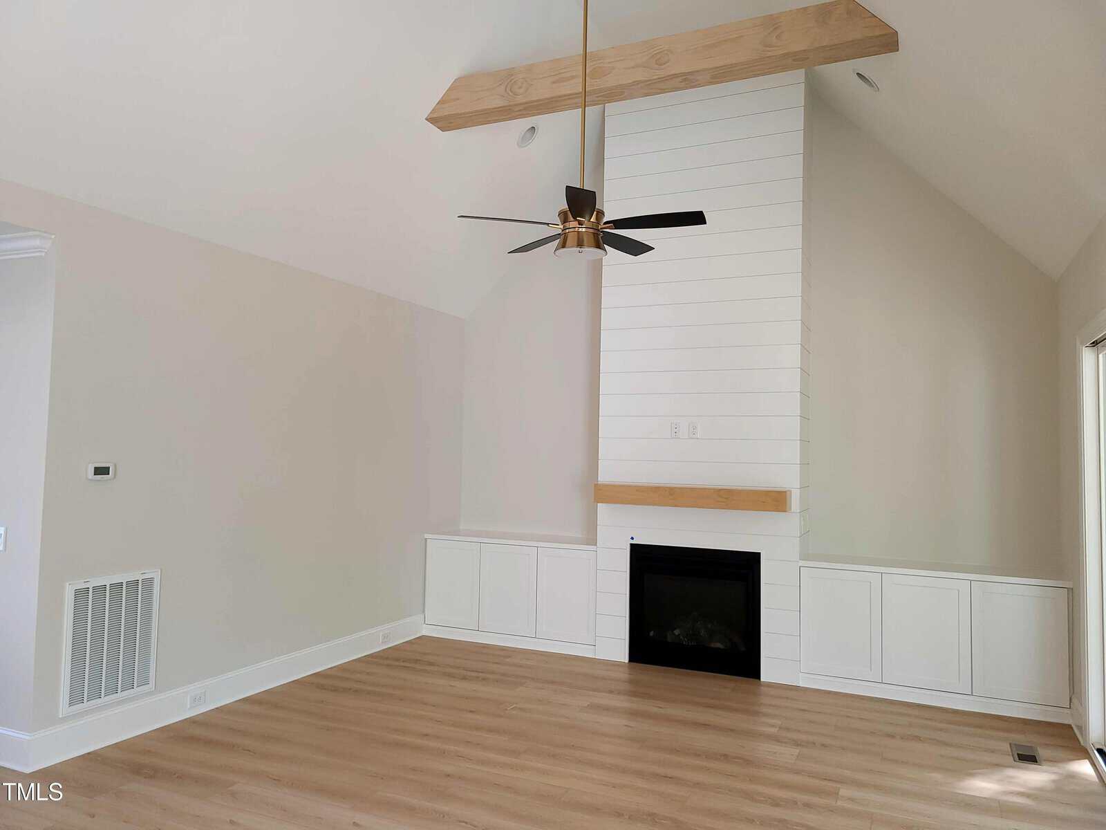 Modern living room with high vaulted ceiling, featuring a white shiplap accent wall and a wooden beam. There's a black fireplace centered below the shiplap, flanked by white cabinets. A ceiling fan hangs above, and the floor is light-colored wood.