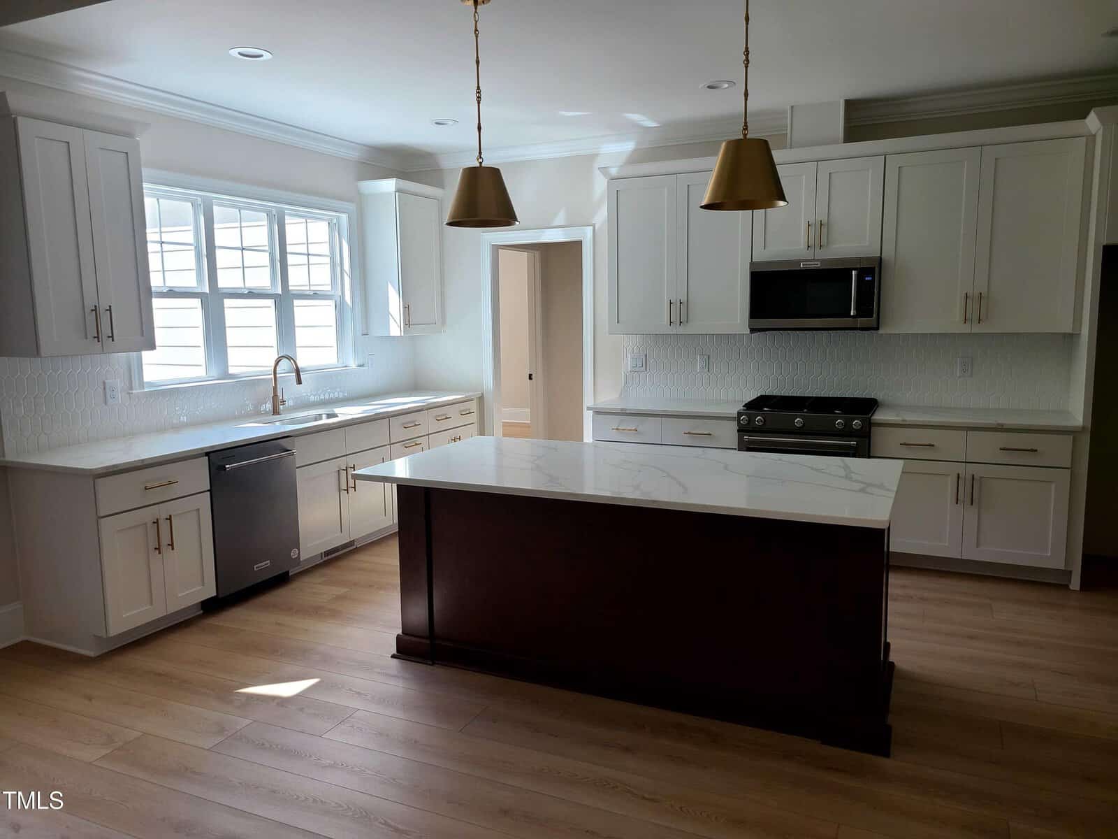 Modern kitchen with white cabinets, marble countertops, and a central island. Two pendant lights hang above the island. Stainless steel appliances, including an oven and dishwasher, are present. Large windows provide natural light.