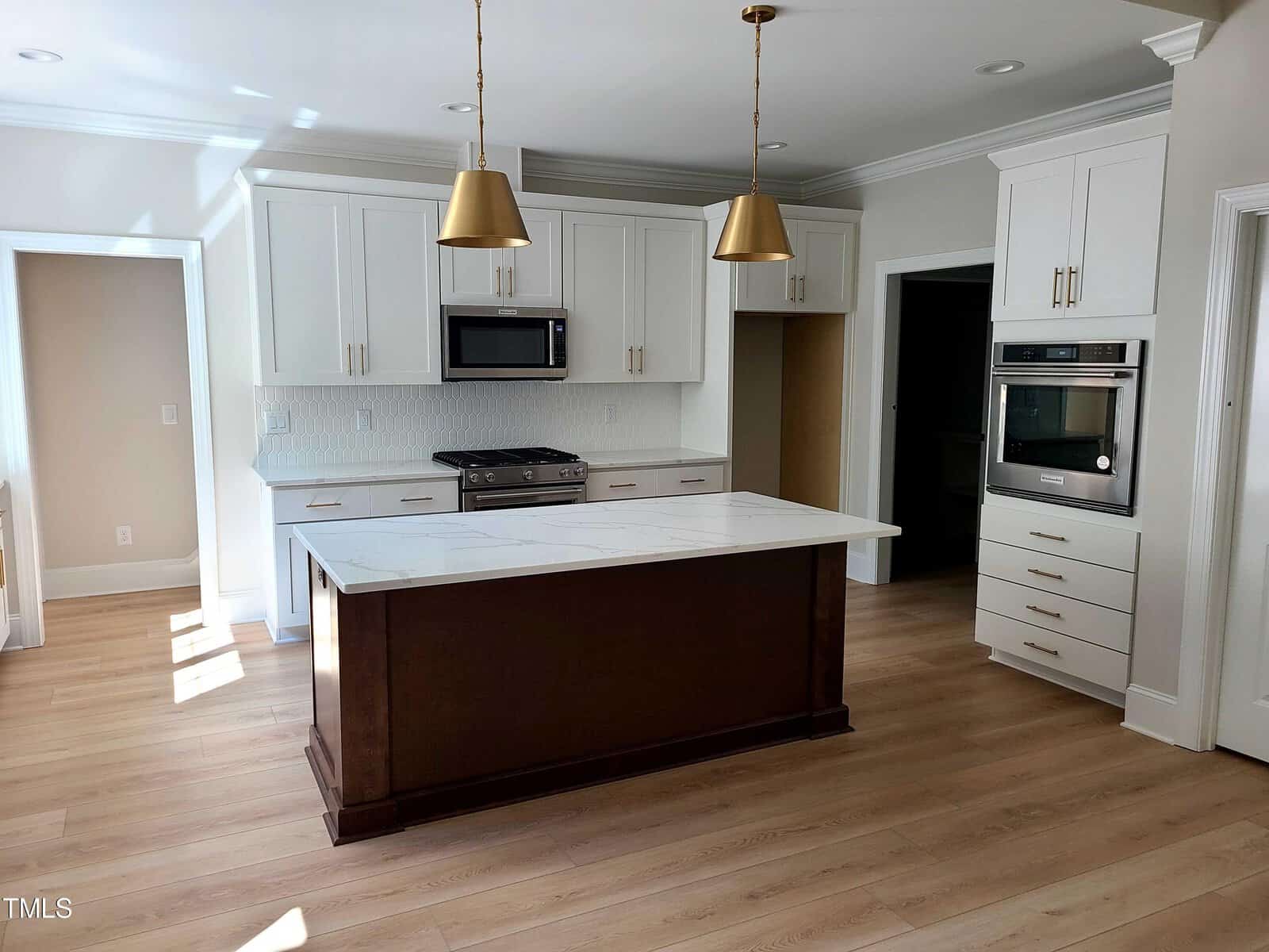 A modern kitchen with white cabinets, stainless steel appliances, a central island with a dark wooden base, and two gold pendant lights. The room has light wood flooring and receives natural light from an unseen window.