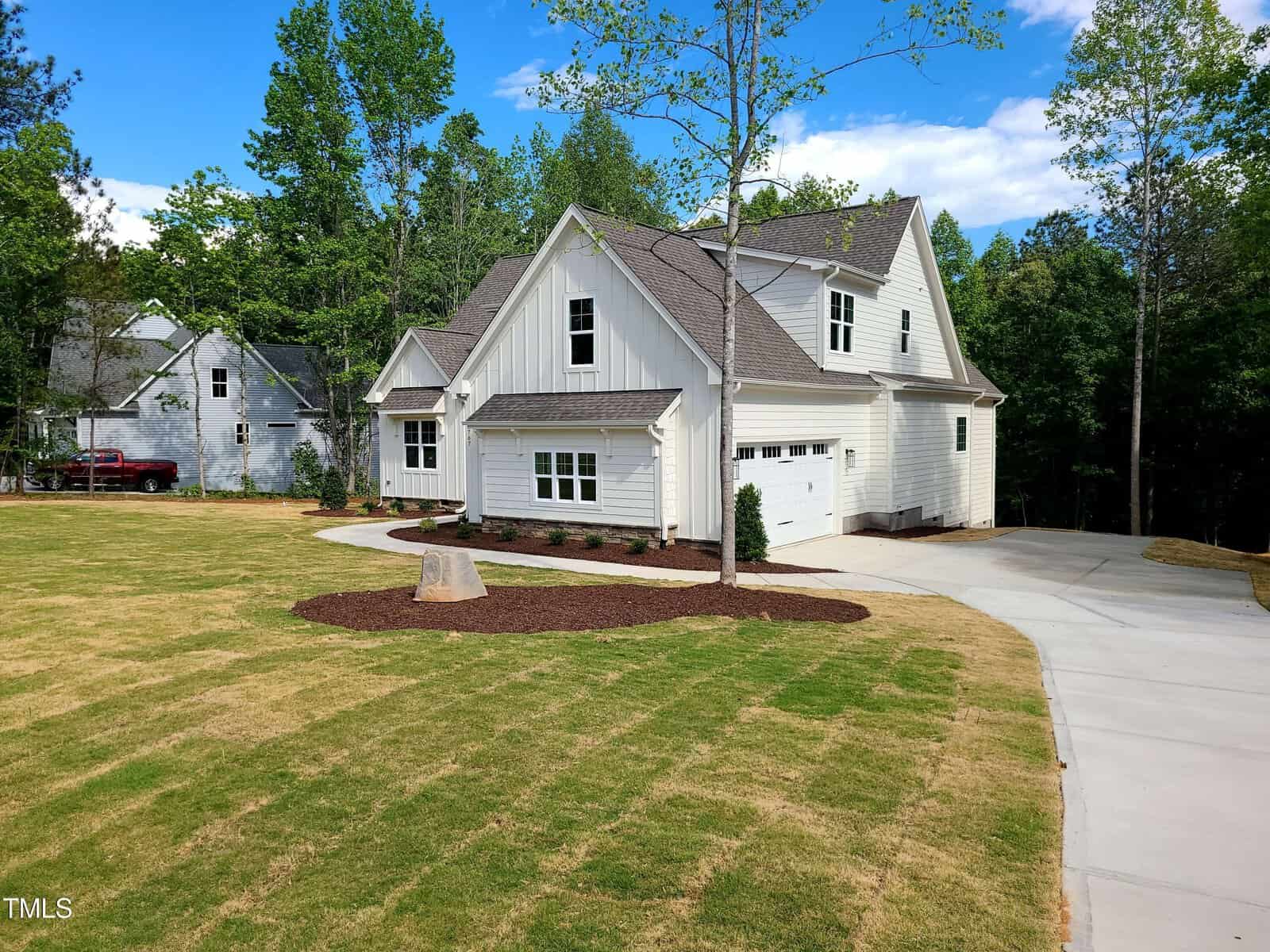 A modern white house with a two-car garage sits beside a freshly landscaped yard. The driveway curves to the entrance, surrounded by lush green trees and a clear blue sky. Another house is visible in the background.