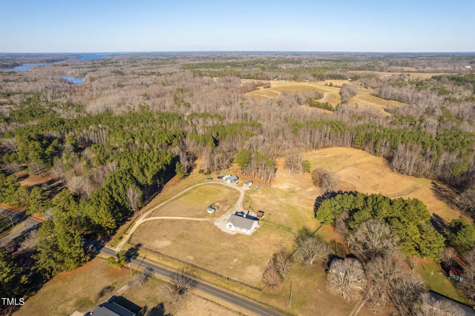 Aerial view of a sprawling rural property featuring a house with a white fence surrounded by a large, manicured lawn, trees, and open land under a clear sky.