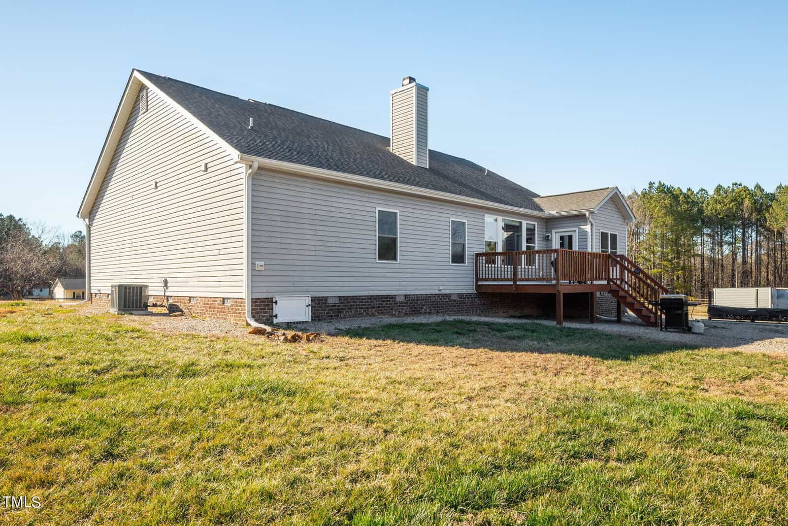 A suburban single-story house with beige siding, featuring a large wooden deck and a grassy backyard under a clear sky.
